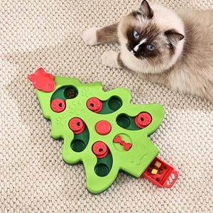 May include: A green, Christmas tree-shaped cat puzzle toy with red details. The toy features compartments and sliding elements for treats. A cat with blue eyes and brown markings is visible in the background.