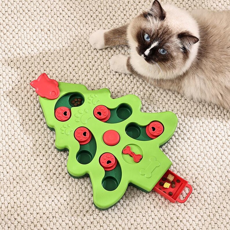May include: A green, Christmas tree-shaped cat puzzle toy with red details. The toy features compartments and sliding elements for treats. A cat with blue eyes and brown markings is visible in the background.