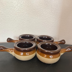 May include: A set of four ceramic soup crocks with handles. Each bowl features a two-tone design with a dark brown glazed rim and handle, transitioning to a lighter tan base. The interior of each bowl is finished in a matching dark brown glaze.