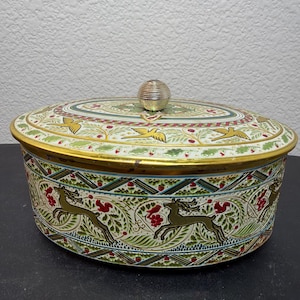 May include: An oval metal storage tin with a removable lid featuring a gold-toned knob. The exterior is decorated with a repeating pattern of green leaves, red berries, and gold-colored deer silhouettes against a cream-colored background.