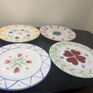 May include: Four round ceramic plates with hand-painted designs. One features yellow diamonds and blue flowers, another has red sunburst lines, one displays four pink tulips with blue triangles, and the last shows a large red flower with green leaves.