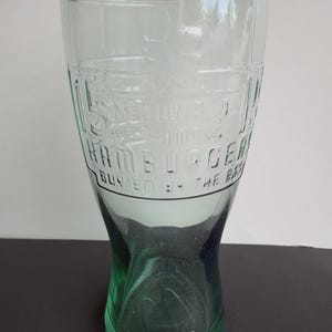 May include: A clear glass tumbler with a tapered base and a wider rim. The glass features embossed text that reads "HAMBURGER" and "BUY EM BY THE BAG" along with a faint graphic of a burger. The base of the glass has a distinct green tint.