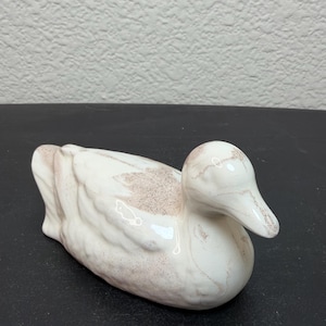 May include: A ceramic duck figurine with a glossy white finish and subtle, light brown speckled detailing across the wings, head, and beak. The duck is depicted in a resting position with its head turned slightly to the right, showcasing smooth curves.
