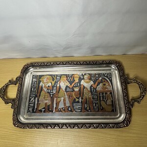 May include: A rectangular metal serving tray with ornate handles and a decorative border. The center features a colorful Egyptian-themed relief depicting three figures, a bull, and hieroglyphics against a dark background, finished in metallic tones.