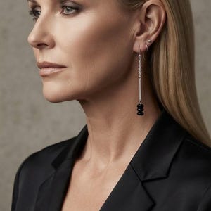 May include: A long drop earring featuring a silver-toned hook, a vertical silver-toned bar, and three small black spherical beads at the base. The earring hangs from the earlobe, complemented by a small silver-toned hoop and a tiny stud earring.