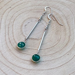 May include: A pair of silver dangle earrings with a modern design. Each earring features a faceted, round, emerald-green gemstone at the bottom, connected to a straight silver bar. The top has a beaded accent and a hook closure. The earrings are displayed on a light wood surface.
