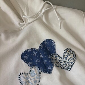 May include: White hoodie with three heart appliqués in shades of blue floral fabric. The hearts are stitched onto the front of the sweatshirt. The hoodie has a drawstring and a front pocket.