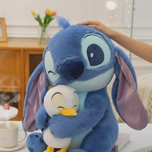 May include: A large, plush blue Stitch doll winks while hugging a small, white and yellow plush duck. The Stitch doll has large ears with pink insides. A hand gently rests on Stitch's head. The background includes a table, a coffee cup, and a vase of flowers.