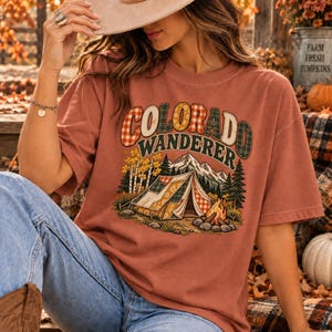 Puede incluir: Una camiseta de manga corta color óxido con un estampado gráfico que incluye el texto COLORADO WANDERER. El diseño muestra una tienda de campaña, una fogata, pinos y montañas, todo en un estilo de patchwork con patrones de cuadros y flores.