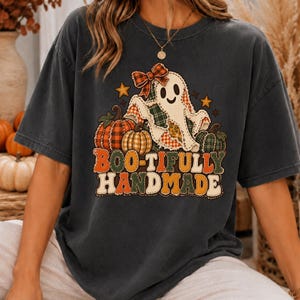 May include: A charcoal grey short-sleeve t-shirt featuring a graphic of a ghost wearing a plaid bow, surrounded by pumpkins and stars. The text BOO-TIFULLY HANDMADE is printed in a multi-coloured, stitched-style font below the ghost illustration.