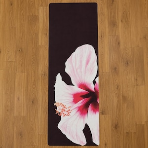 May include: A rectangular yoga mat featuring a solid black background with a large, detailed graphic of a white and pink hibiscus flower. The flower petals display deep magenta centers and prominent orange-tipped stamens extending to the left side.