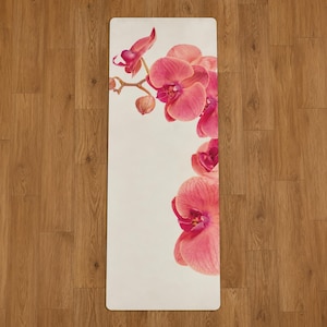 May include: A rectangular yoga mat with a light cream-colored background featuring a vibrant floral print of pink and magenta orchids. The orchid stems and blossoms are arranged along the top left and right edges of the mat surface.