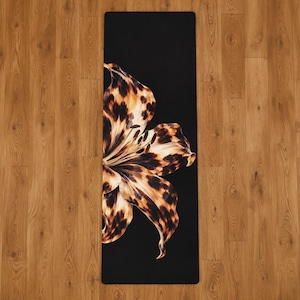 May include: A rectangular yoga mat featuring a solid black background with a large, detailed lily flower graphic. The petals of the lily are patterned with a brown and black leopard print design, extending from the left side across the center.