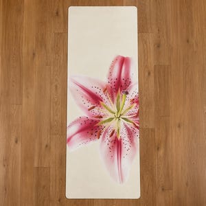 Puede incluir: Una esterilla de yoga rectangular con fondo color crema que presenta una gran flor de lirio rosa centrada. La flor tiene motas oscuras prominentes en sus pétalos, un centro verde claro y estambres marrones visibles que salen del medio.