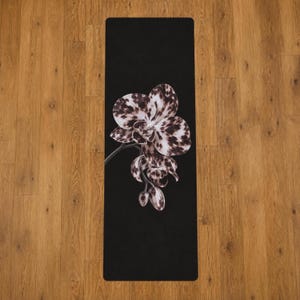 May include: A rectangular yoga mat with a solid black background featuring a large, detailed illustration of an orchid flower. The orchid petals display a mottled, spotted pattern in shades of white, grey, and dark brown, with a thin stem.