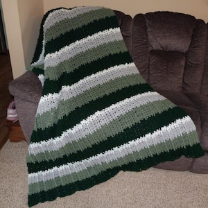 May include: A handmade crochet blanket draped over a brown sofa. The blanket features horizontal stripes in shades of green, gray, and white. The textured pattern adds visual interest to the cozy home decor item.