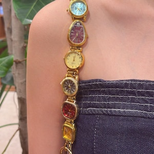 May include: A decorative strap constructed from a series of gold-toned wristwatches with various dial colors including light blue, deep red, yellow, and black, connected in a vertical line to form a unique accessory for a dark blue denim garment.