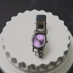 May include: A silver-toned metal clip featuring an integrated oval-shaped wristwatch with a vibrant purple dial. The watch face displays silver hour markers and hands, with a metallic silver casing that connects directly to the spring-loaded clip mechanism.