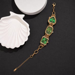 May include: A gold-toned bracelet watch featuring three distinct watch faces with emerald green dials. The faces are heart-shaped, rectangular, and square, connected by gold-toned links. The square face is accented with small clear stones.