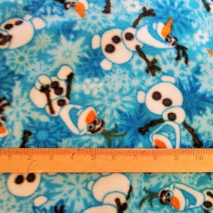 May include: Blue fleece fabric with a repeating pattern of white Olaf snowmen from the movie Frozen. The fabric is measured with a wooden ruler.