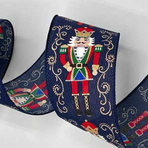 May include: Blue ribbon with a repeating pattern of a nutcracker in a red and green uniform with a gold crown and gold trim. The background is a dark blue with gold swirls.