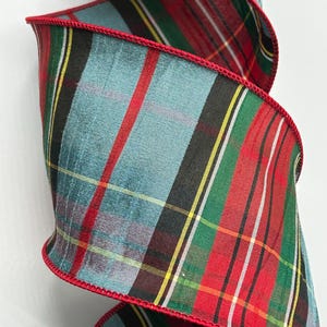 May include: A close-up of a decorative ribbon with a plaid pattern. The ribbon features a teal, red, green, black, and yellow design with a red stitched edge. The ribbon is curled and appears to be made of fabric.