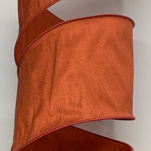 May include: Close-up of a wide, spiraling ribbon in a warm, burnt orange color. The ribbon has a textured appearance and is edged with a darker, reddish-brown trim. The ribbon's design suggests it could be used for decorative purposes.