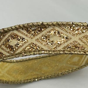 Puede incluir: Primer plano de una cinta dorada con un patr&oacute;n de diamantes. La cinta tiene una base de color crema con detalles de purpurina dorada dentro de las formas de diamante. Los bordes est&aacute;n adornados con hilo dorado, creando un adorno decorativo. Ideal para manualidades o envolver regalos.