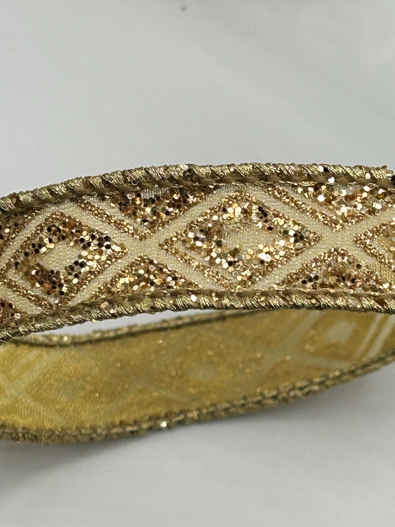 Puede incluir: Primer plano de una cinta dorada con un patr&oacute;n de diamantes. La cinta tiene una base de color crema con detalles de purpurina dorada dentro de las formas de diamante. Los bordes est&aacute;n adornados con hilo dorado, creando un adorno decorativo. Ideal para manualidades o envolver regalos.