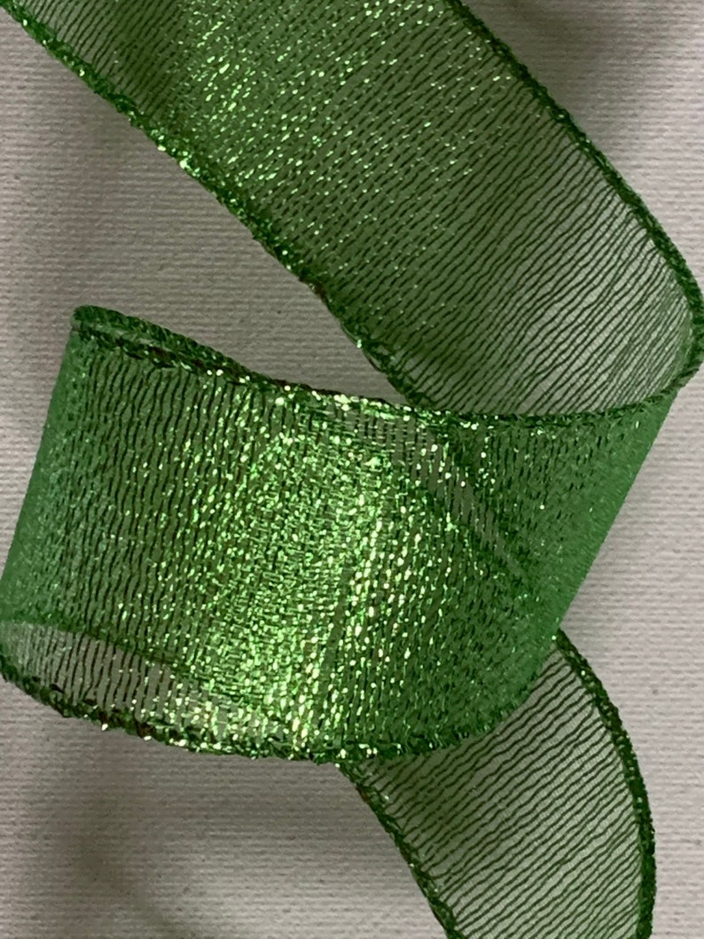 May include: Close-up of a vibrant green ribbon with a shimmering, glittery texture. The ribbon is curled, showcasing its width and the intricate weave of the material. The edges are neatly finished, adding to its decorative appeal.