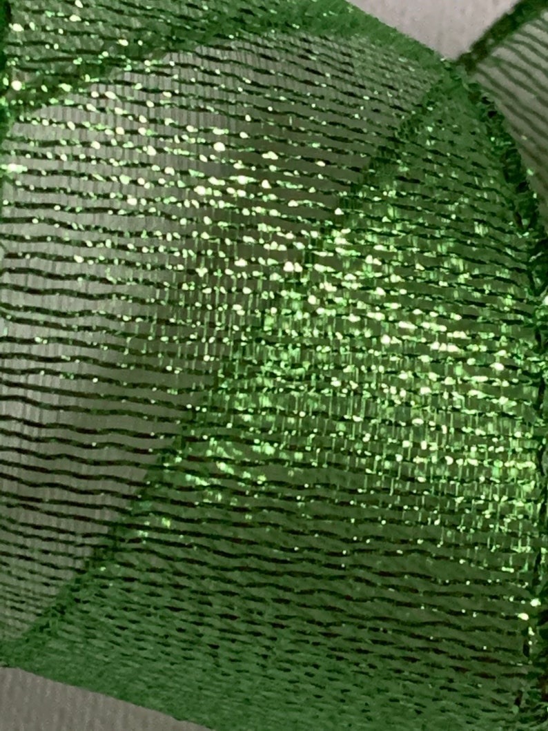 May include: Close-up of a shimmering green mesh ribbon. The ribbon has a textured, woven appearance with horizontal lines and a metallic sheen. The material reflects light, creating a sparkling effect, suitable for crafts and decorations.