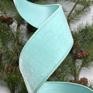 May include: A wide, light blue velvet ribbon is spiraling in the image. The ribbon has a soft, plush texture and is edged with a stitched border. Pine branches and pine cones are in the background, suggesting a holiday or seasonal theme.