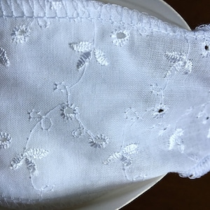 May include: Close-up of a white fabric with embroidered floral and leaf patterns. The fabric has a delicate, airy appearance, with small cut-out details. The edge is finished with a scalloped trim, adding a touch of elegance to the design.