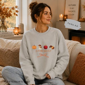 Könnte beinhalten: Ein aschfarbenes Sweatshirt mit Rundhalsausschnitt und einem Grafikdruck aus einem orangefarbenen Kürbis, braunen Pilzen, einem roten Apfel und einer Eichel. Unter den Abbildungen steht der Text Autumn Harvest sowie eine rot-weiß karierte Schleife.
