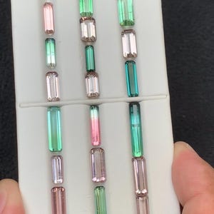 Puede incluir: Una colección de diecisiete piedras preciosas de turmalina rectangulares, con corte baguette, dispuestas en tres columnas verticales sobre una bandeja de presentación blanca. Las piedras presentan colores translúcidos, incluyendo tonos rosa, melocotón y verde.