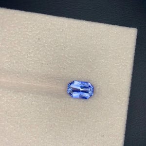 Könnte beinhalten: Ein rechteckiger, facettierter, hellblauer Edelstein. Der Edelstein ist vor einem weißen, strukturierten Hintergrund platziert. Die Facetten des Edelsteins reflektieren das Licht und erzeugen einen funkelnden Effekt. Der Edelstein hat einen lebendigen Blauton.