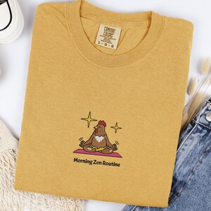 Puede incluir: Camiseta de color amarillo mostaza con cuello redondo. La camiseta presenta un diseño bordado de un pollo meditando sobre una esterilla de yoga rosa, con el texto "Morning Zen Routine" debajo. La camiseta tiene una etiqueta que dice "Comfort Colors".