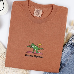 Könnte beinhalten: Ein rostfarbenes T-Shirt mit Rundhalsausschnitt. Das Shirt zeigt eine Grafik einer grünen Frosches in einer Yoga-Pose auf einer rosa Matte, mit dem Text "Hop Into Alignment" darunter. Das Shirt hat ein Etikett mit der Aufschrift "Comfort Colors".