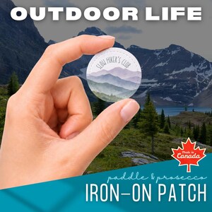 Op de afbeelding: Een ronde opstrijkbare patch met een aquarelillustratie van een berglandschap en de tekst "SLOW HIKER'S CLUB" in een donker lettertype. De patch wordt getoond tegen een achtergrond met een bergketen, een meer en de tekst "OUTDOOR LIFE".