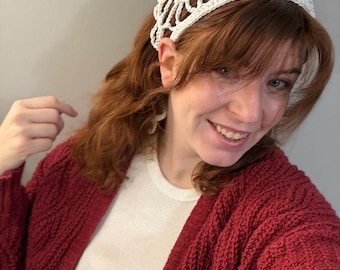 Könnte beinhalten: Ein weißes gehäkeltes Haaraccessoire mit einem durchbrochenen Netzmuster. Die Kopfbedeckung besteht aus Garn und ist so konzipiert, dass sie über dem Haar getragen und mit Bändern am Hinterkopf befestigt wird, um den Halt zu gewährleisten.
