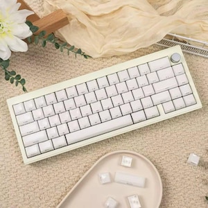 Op de afbeelding: Een crèmekleurig mechanisch toetsenbord met witte toetsen en een ronde zilveren knop in de rechterbovenhoek. Het toetsenbord heeft een standaard QWERTY-indeling met witte letters op elke toets. Een klein schaaltje met extra toetsen ligt ernaast.