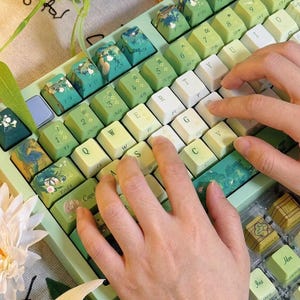May include: A mechanical keyboard with a light green frame and custom keycaps featuring botanical illustrations, floral patterns, and landscape art. The keys are a mix of light green, white, and dark green, with some keys displaying printed text and symbols.