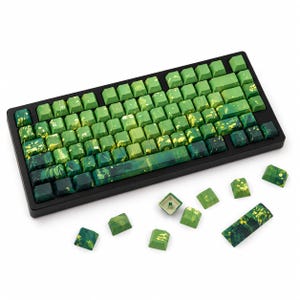 Pode incluir: Um teclado mecânico com uma estrutura preta e um conjunto de teclas personalizadas com um padrão de salpicos de tinta marmoreada em verde e amarelo. Várias teclas soltas a condizer estão espalhadas à frente do teclado, exibindo o mesmo esquema de cores verde e amarelo e um acabamento texturado.