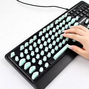 May include: A black computer keyboard featuring custom light mint green keycaps shaped like fluffy clouds. Each individual keycap has a cloud-like silhouette, replacing standard square keys, while the space bar remains a smooth rectangular shape.