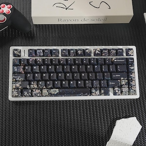 Könnte beinhalten: Eine mechanische Tastatur mit weißem Rahmen und dunkelblauen Tastenkappen, die mit einem detaillierten Blumenmuster in Blau-, Creme- und Grüntönen verziert sind. Die Tasten zeigen Standard-QWERTY-Buchstaben und Symbole in Weiß, in kompakter Anordnung.