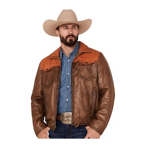 May include: A beige felt cowboy hat paired with a brown leather jacket featuring a contrasting tan suede yoke across the shoulders. The jacket includes a front zipper, two chest pockets with flaps, and long sleeves. A blue button-down shirt is worn underneath.