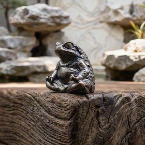 Peut inclure: Une petite figurine en métal sombre représentant un crapaud assis en position de méditation, les jambes croisées. Le crapaud présente un dos texturé et bosselé ainsi qu'un ventre lisse et arrondi, avec ses membres antérieurs en position.