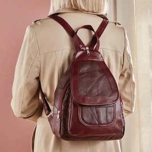 May include: A burgundy leather backpack with a flap closure and a front pocket. The backpack has adjustable straps and a top handle. The leather has a patchwork design. The person is wearing a beige trench coat.