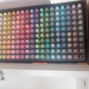 May include: A large rectangular black storage box containing a vast collection of markers arranged in a grid. The markers are organised by colour, displaying a full spectrum including shades of yellow, orange, pink, purple, green, blue, and grey.