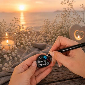 Peut inclure: Une pierre noire lisse avec l'inscription "In loving memory" peinte en blanc, ornée d'une petite fleur bleue. Un marqueur noir est utilisé pour ajouter des détails sur la pierre, qui est maintenue en place pendant sa décoration.